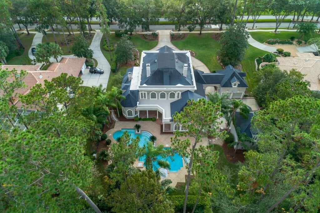 Arial drone shot of a large white house with blue roof surrounded by trees. - HomeJab