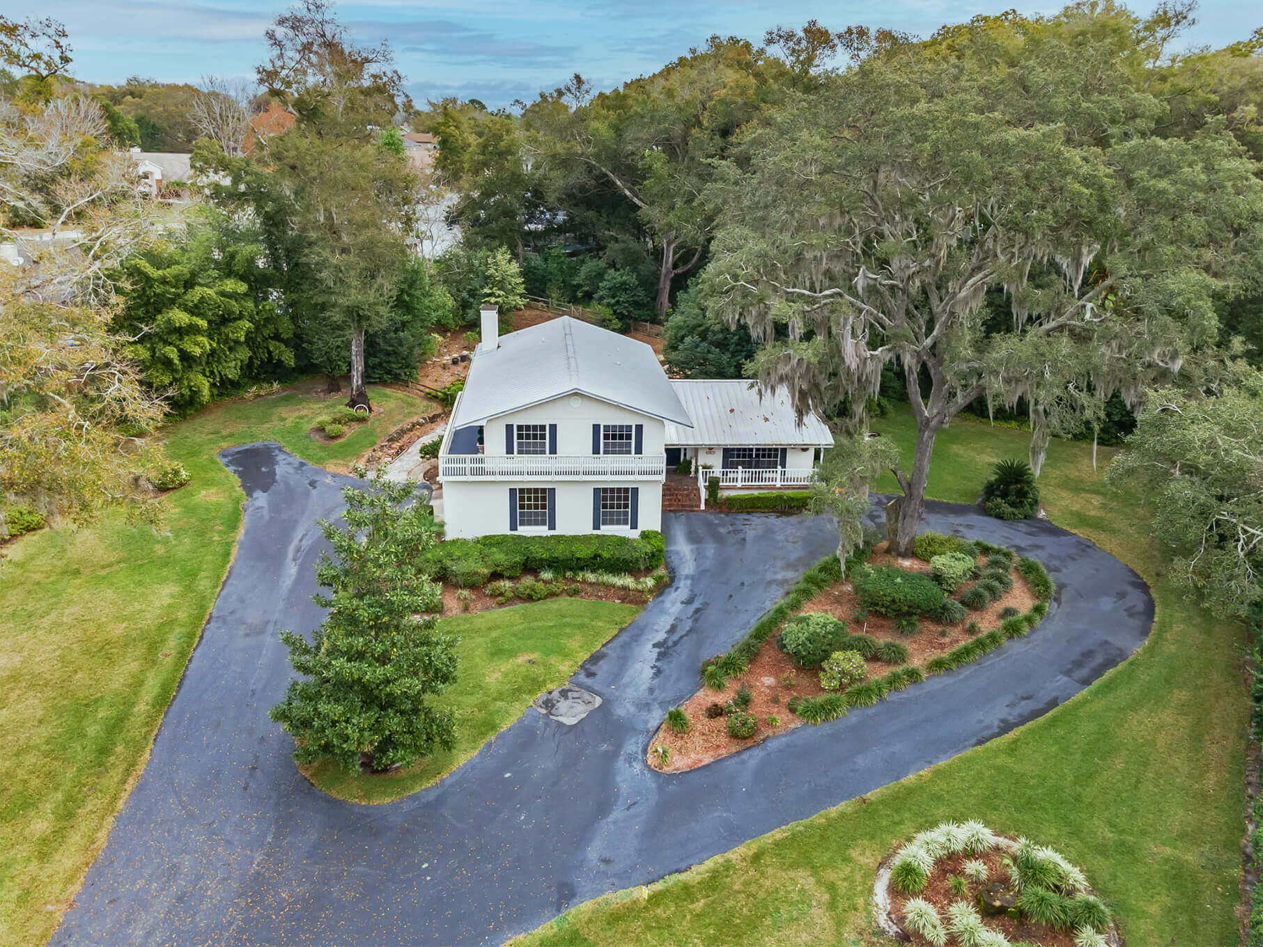 Arial drone photograph of a two story home surrounded by tall trees and a winding driveway.