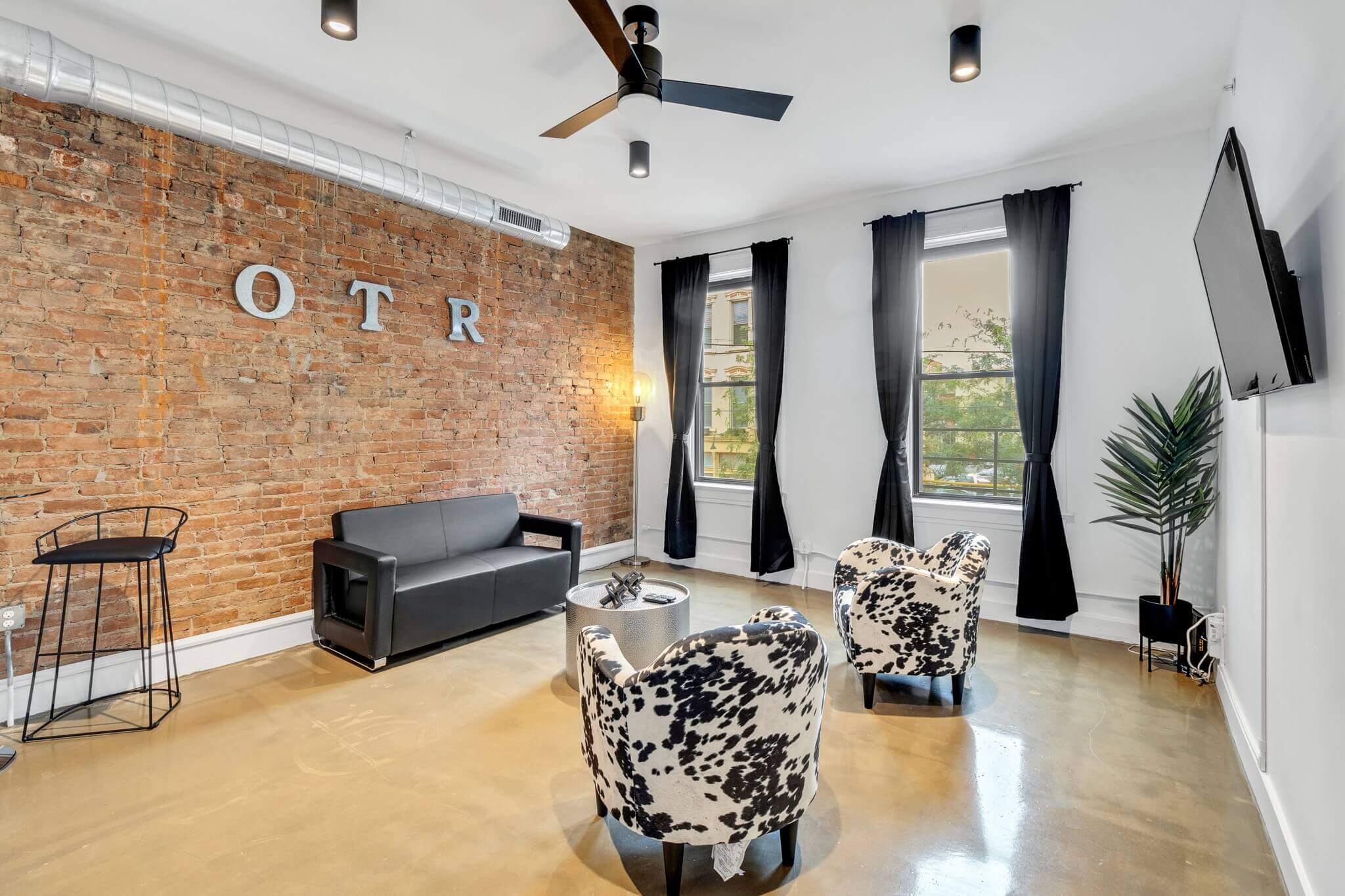 Living room with white walls and a brick accent wall with black curtains and ceiling fan.